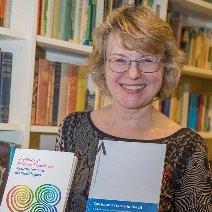 Standing in front of a bookcase, Professor Bettina Schmidt smiles towards the camera while holding up two of her own works: The Study of Religious Experience – Approaches and Methodologies; also Spirits and Trance in Brazil. 