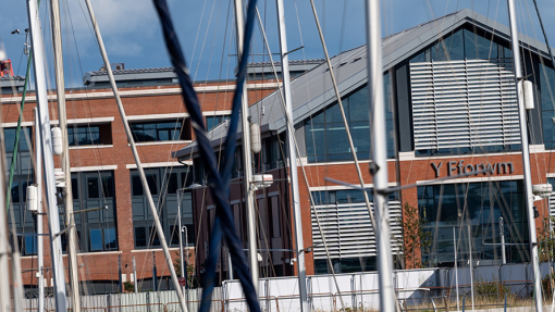Y Fforwm Library, a large building with pillars of red brick and much glass. The masts of yachts are visible in the foreground.