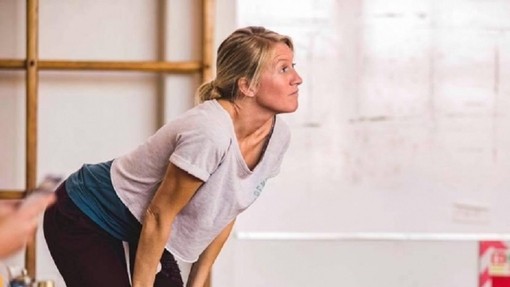In a rehearsal room Angharad Lee leans forwards, hands on her knees, as she observes something off-camera.
