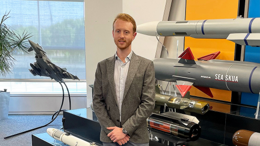 Wearing a grey jacket over an open-necked shirt, Max Hart stands in front of a display of missile casings, including a Sea Skua.