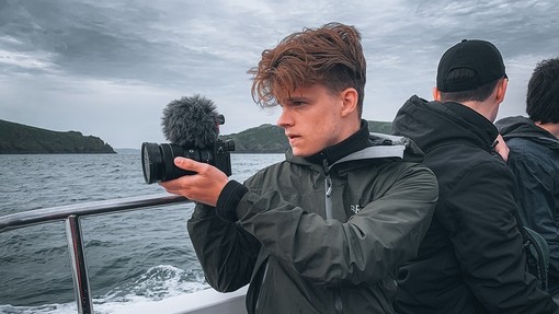 On a grey day, Joshua Knight sits near the stern of a boat and turns towards the sea, holding up a camera with a microphone attachment.
