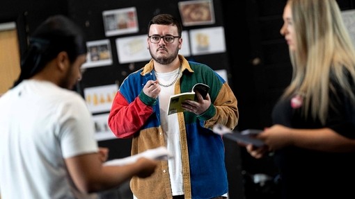 William Kingshott stands in a rehearsal room; he holds a playscript open in one hand and has a pencil in the other; his eyes are focused on another student.