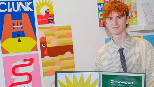 Evan Elias smiles slightly towards the camera as he stands by an exhibition of his illustrations.