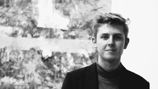 A black-and-white photo of Owain Sparnon wearing a black coat over a polo-neck; he is standing in front of an abstract, textured artwork.