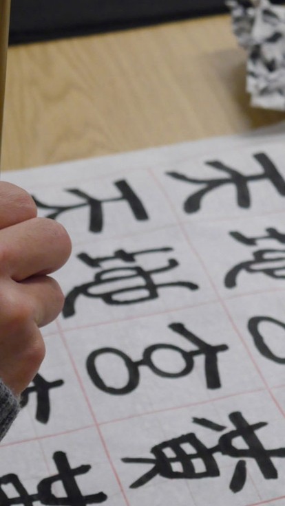 A hand holding a wooden calligraphy brush over a page of Chinese characters.