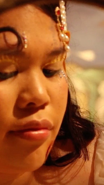 The face of a young woman, her hair and forehead decorated with jewellery and her eyes closed, is seen in soft yellow light through the hollow centre of a golden disc. 