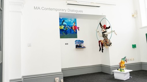 A large carnival doll hanging in a hoop from the ceiling and other exhibits beneath the words MA Contemporary Dialogues.
