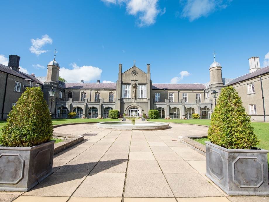 Picture of outside Lampeter Campus, grey brick building with grey flagstone walkway with a water fountain in the centre and two green bushes in grey square plant pots on a sunny day