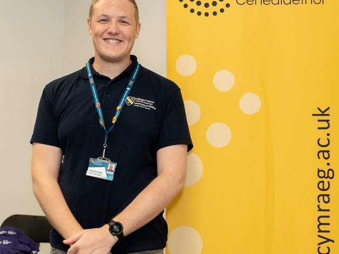 Staff member standing in front of a yellow Coleg Cymraeg banner