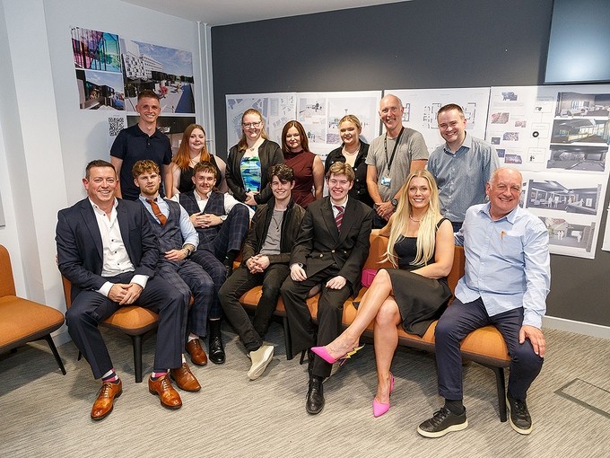 A group of students and staff either stand or sit and smile cheerfully for a group photo; the students have dressed up for the occasion in evening wear; behind them you can see displays of architectural plans.