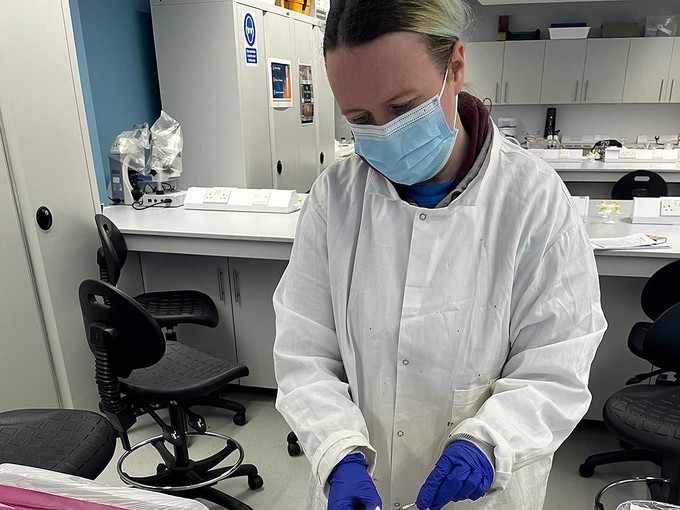 Wearing a facemask and white labcoat, Rebecca Macfarlane looks down, focusing on her work; she is in a room with long white workbenches and cupboards with hazard signs.