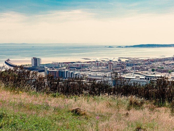 A view of SA1 and Swansea Bay on a sunny day; a grassy hillside occupies the foreground; beyond that the marina, the beach, the water of the bay and the headland of the Mumbles are visible.