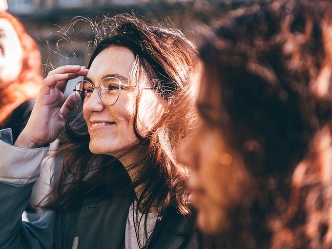 Outside, standing in a group of young people, a young woman in glasses smiles as she brushes back a lock of hair.