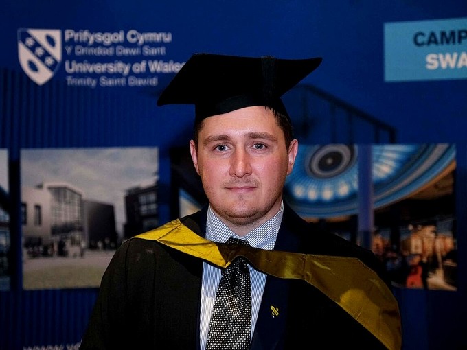 Jon Langstone wearing an academic gown, golden hood, and mortarboard hat.