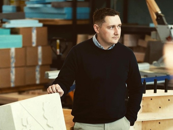 Jon Langstone standing with one hand in his pocket and the other resting on the corner of a metre-high coat of arms in newly carved stone.