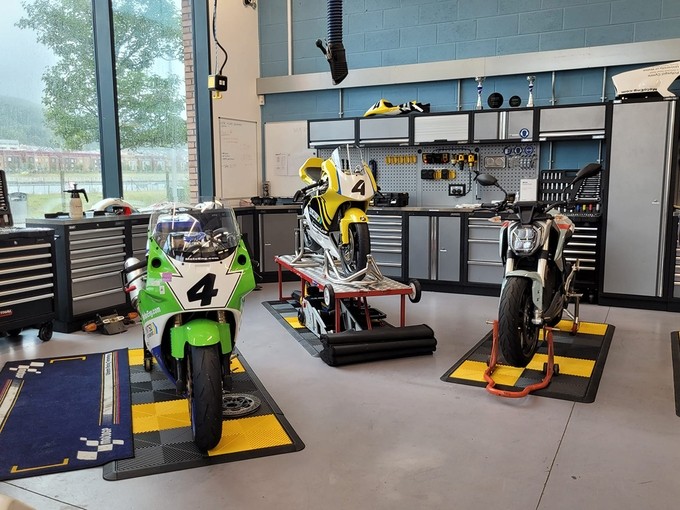 Three motorbikes, including two in racing colours, stand in a workshop with large windows.