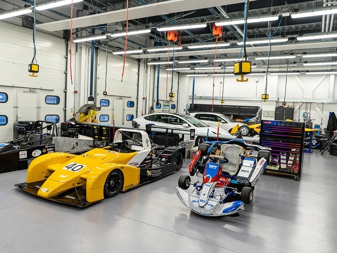 A large well-lit garage with three racing cars in the foreground at different stages of assembly.