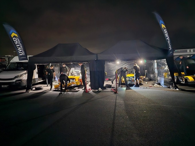Students work at night on a pair of racing cars in a parking lot; each car is sheltered under a pop-up black gazebo.