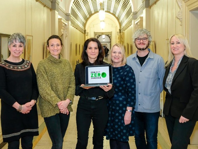 Agnes Olah smiles as she holds up a laptop with Project Zero Swansea on the screen; she is flanked by representatives of Swansea Council and UWTSD.