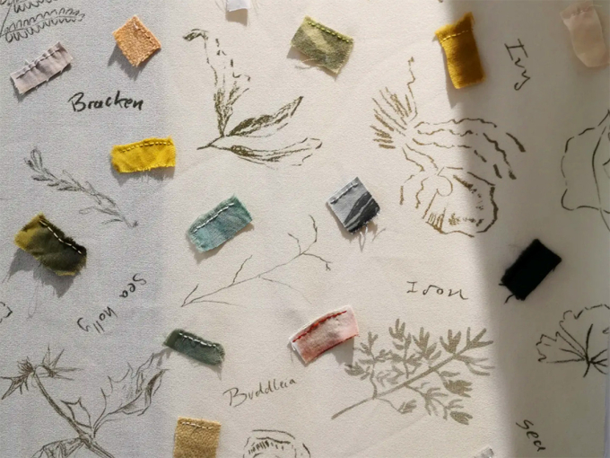 Small fabric cuttings sewn onto a cream-coloured cloth; the cloth is decorated with botanical prints and handwritten labels, including: buddleia, ivy, bracken, sea holly.