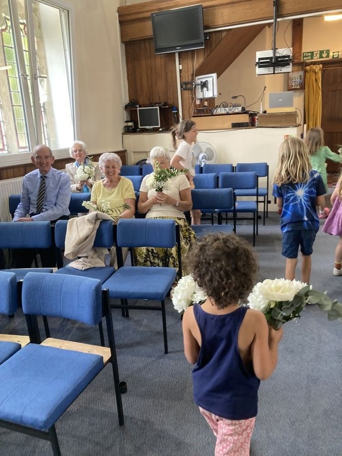 Children carry bouquets into a hall with seated older people. 