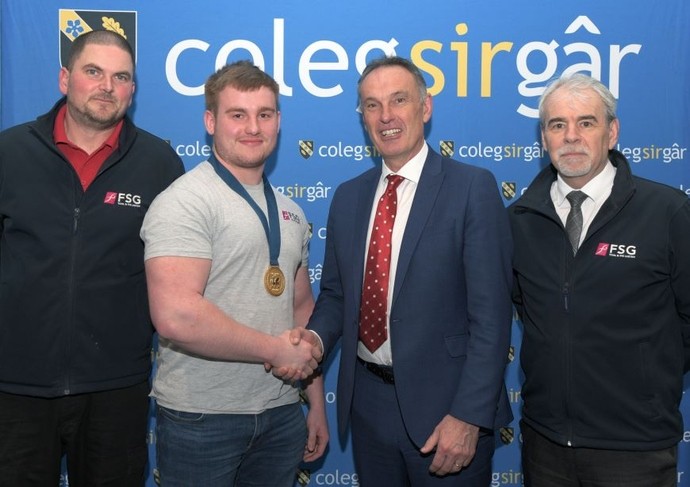 Student Kian Lloyd proudly wearing a gold medal shakes hands with Deputy Vice Chancellor Dylan Jones, celebrating his competition win; two representatives from FSG Tool and Die Limited are also present.