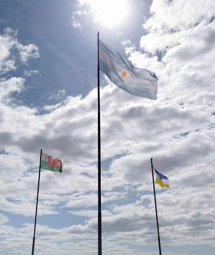 Three flags are raised on masts in front of a sunny backdrop.