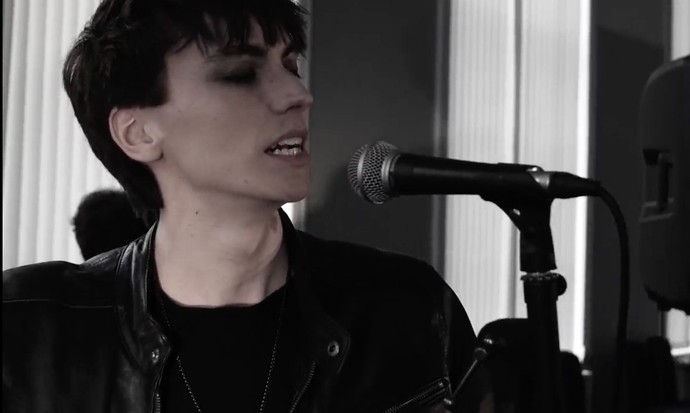 A desatured photo showing a young man in a black leather jacket with black mascara and black hair singing into a microphone. 