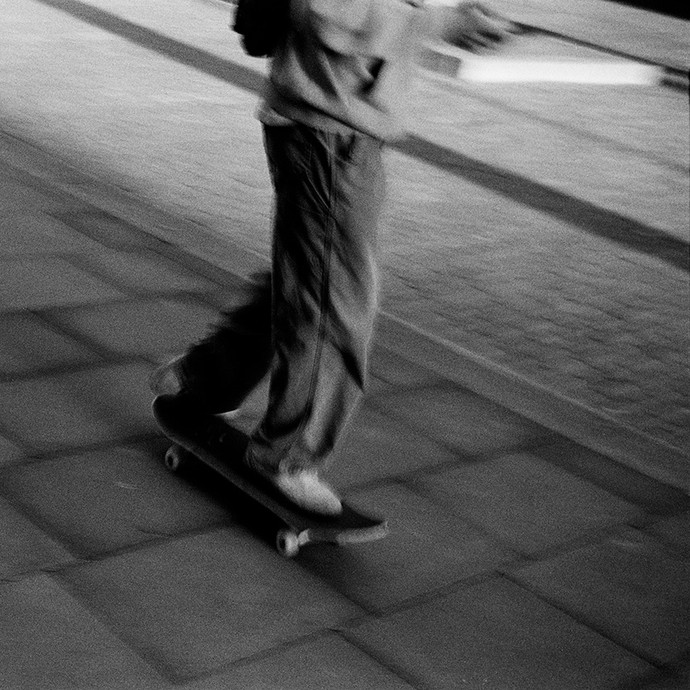 A black-and-white photograph showing someone with a rucksack, phone, and baggy clothes skateboarding along a pavement; the face isn't shown. 