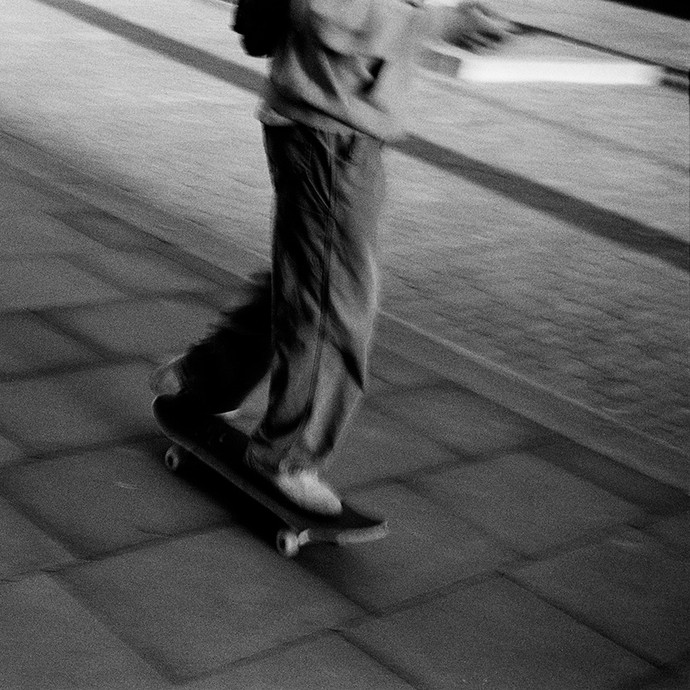 A black-and-white photograph showing someone with a rucksack, phone, and baggy clothes skateboarding along a pavement; the face isn't shown.