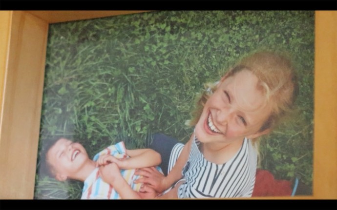 A blonde girl of around eleven looks up smiling towards the camera; a younger boy lies laughing on the grass as the girl holds him down.