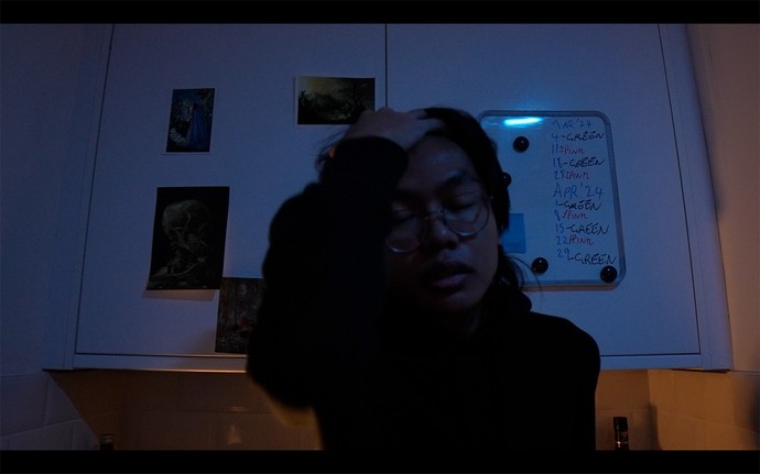 A young man closes his eyes in a dark kitchen, one hand gripping his hair in stress.