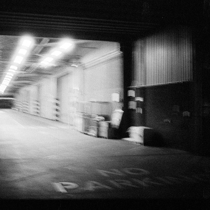 A black-and-white photo showing the entrance to a warehouse at night with the words No Parking on the drive up to the entrance.