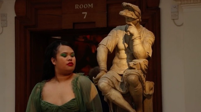 A young Filipino woman with green eyeshadow, a green dress, and red lipstick stands in front of a classical marble statue of a seated man in a muscle cuirass and tunic. 