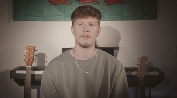 A young man sits looking towards the camera; behind him you can see a keyboard, the heads of two guitars, and a Welsh flag.