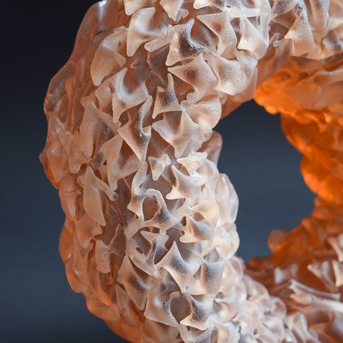 A thick band of pale glass lit with an orange glow in the interior; the surface is composed of hundreds of small folds like cloth. 