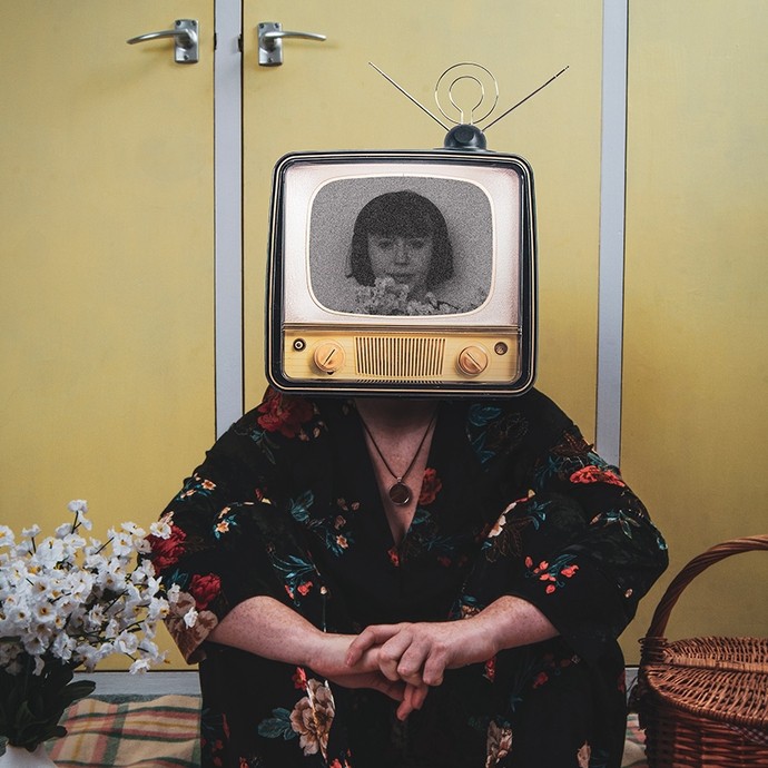 A woman wearing floral pyjamas sits on a tartan rug between a wicker basket, a toy pig and a vase of white flowers; she is wearing a replica of a nineteen sixties TV set on her head; the screen displays a black-and-white photograph of a woman looking out towards the viewer. 