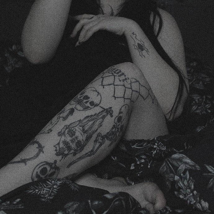 A desaturated photograph showing the body of a woman as she sits on a bed; her right leg displays a series of gothic tattoos including skulls, a spiderweb, and a carved pumpkin.