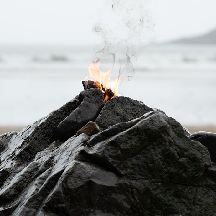 Flames rise form the peak of a craggy reef on a beach; the close-up angle of the photo gives the reef the look of a volcano. 