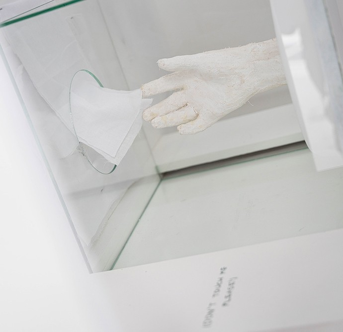 A white plaster hand in a glass box reaching towards a piece of white cloth.