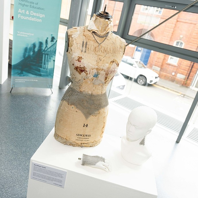 A mannequin on display alongside a mannequin head and hand; its surface has been coated in yellowed, flaking paper;  a small piece of chainmail is visible on all three objects; additionally, the mannequin wears a necklace of metal links. 