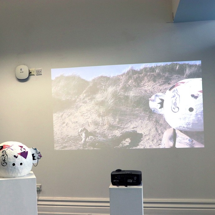 Painted papier-mâché heads displayed on white pedestals; a projection on the wall shows one of the heads resting beside a sand dune. 