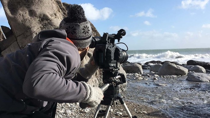 A man crouches by a camera on a tripod as he films white breakers on a rocky shore.