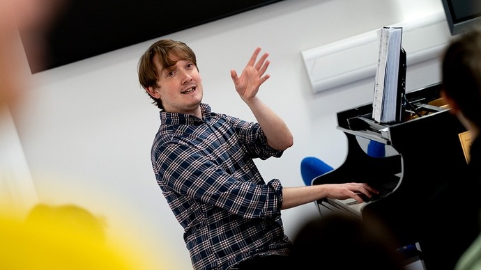 A lecturer stands with one hand on a piano keyboard, the other hand raised in a gesture of emphasis as he explains something to his student audience. 