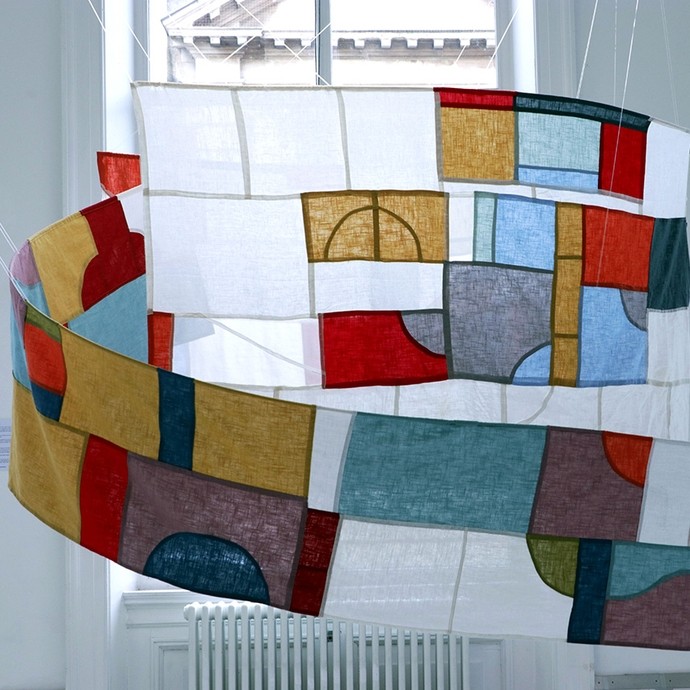 A long banner with white rectangles intercut by other shapes in different colours recalling stained glass; it is situated in front of a window and is suspended in a spiral from the ceiling by translucent cables.