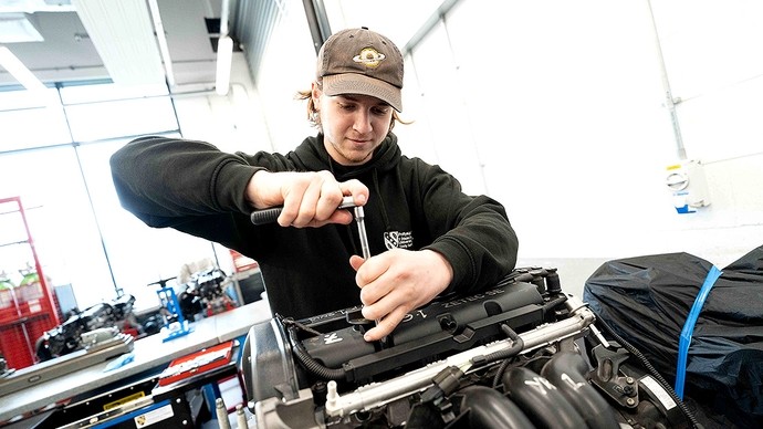 A young man in a black UWTSD hoodie and baseball cap uses a right-angled screwdriver on an engine. 
