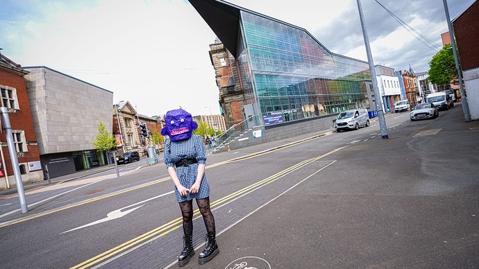 A woman in a check dress and lace-up boots and wearing a purple papier-mâché head stands in front of the modern glass-faced extension to the Alex Building. 