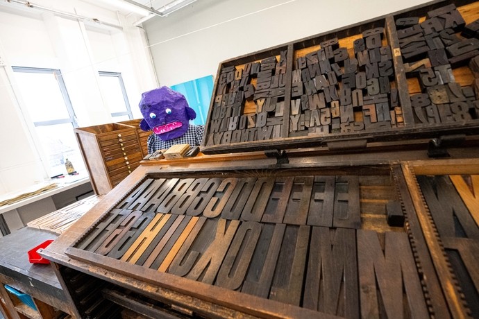 A person wearing a purple papier-mâché mask stands next to large trays full of alphabetic letterpress printing blocks made of wood. 