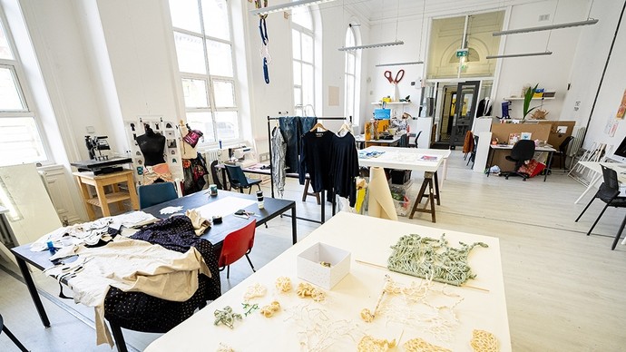 A large white room with floor to ceiling arched windows; tables with scraps of knotwork and textiles, racks of clothes, a sewing machine, and three mannequins indicate the purpose of the studio. 