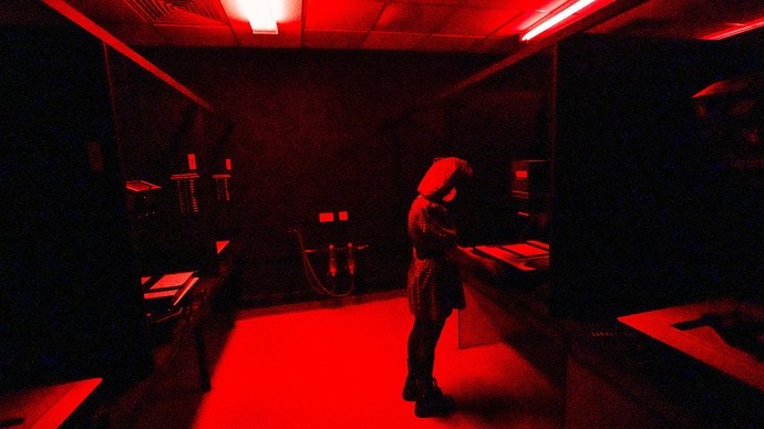 In a darkened room partially lit with red light, a woman wearing a papier-mâché head stands in front of an enlarger on a workbench.